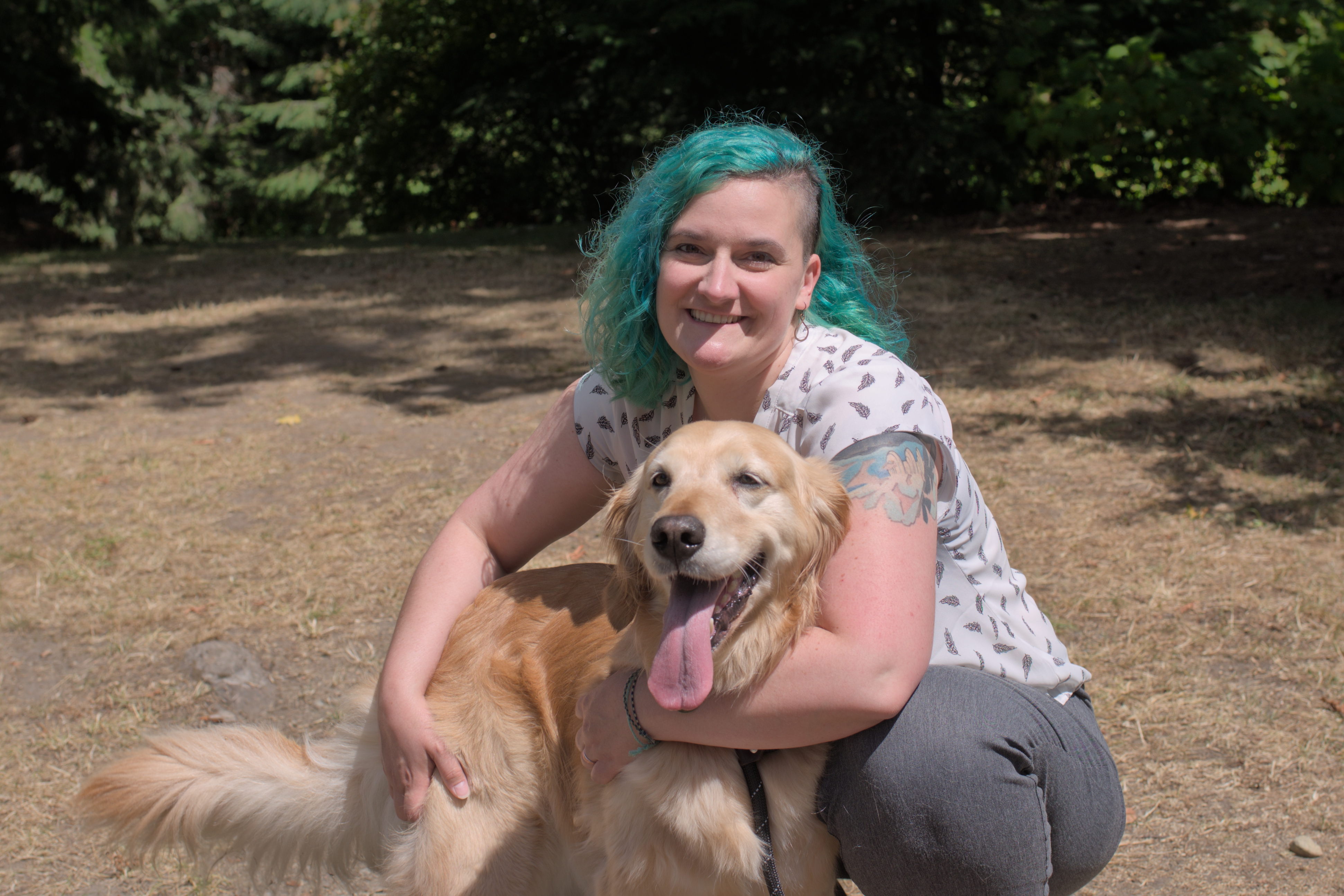 A white woman with teal hair wearing a white shirt kneeling with a golden retriever.